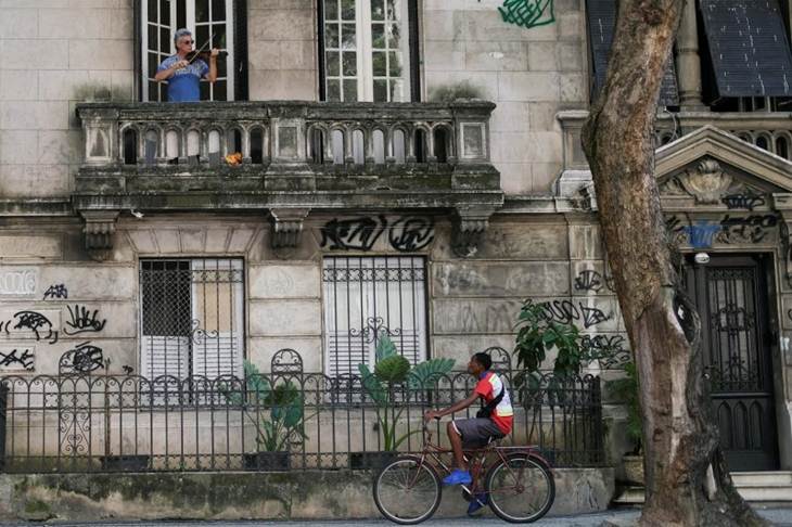 Adelmo Carvalho, 55 tahun, seorang perwakilan penjualan yang biasa bermain di gereja, memainkan biola miliknya di balkon rumahnya saat seorang kurir bersepeda lewat dalam penyebaran penyakit virus korona (COVID-19) di Rio de Janeiro, Brazil, Minggu (29/3/2020). (REUTERS/RICARDO MORAES)